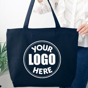 K&ouml;nnte beinhalten: Eine marineblaue Tragetasche mit langen Henkeln. Die Tasche hat ein wei&szlig;es kreisf&ouml;rmiges Logo mit der Aufschrift "YOUR LOGO HERE". Die Tasche wird von einer Person gehalten, die eine wei&szlig;e Bluse und blaue Jeans tr&auml;gt.
