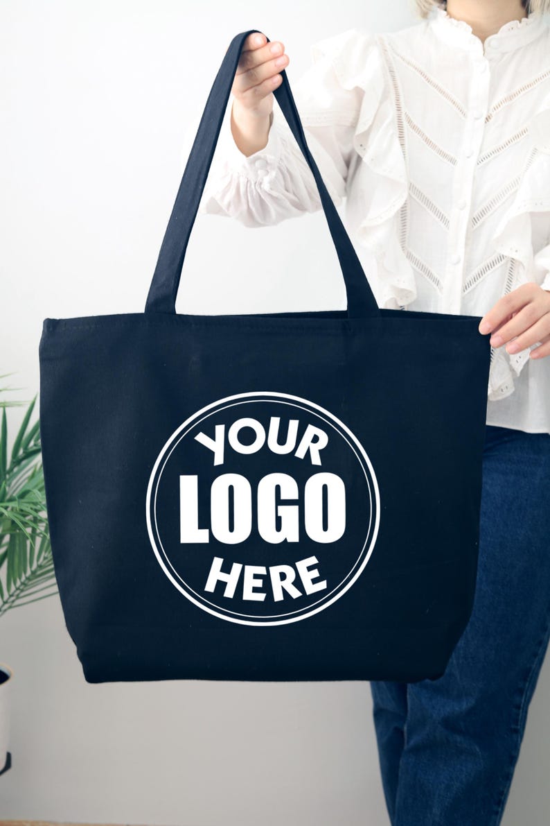 K&ouml;nnte beinhalten: Eine marineblaue Tragetasche mit langen Henkeln. Die Tasche hat ein wei&szlig;es kreisf&ouml;rmiges Logo mit der Aufschrift "YOUR LOGO HERE". Die Tasche wird von einer Person gehalten, die eine wei&szlig;e Bluse und blaue Jeans tr&auml;gt.