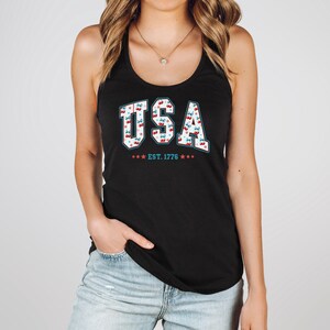 May include: Black tank top with the word "USA" in white, outlined in red, and filled with a cherry pattern. Below the word "USA" is the text "EST. 1776" with red and blue stars. The tank top is worn by a person.