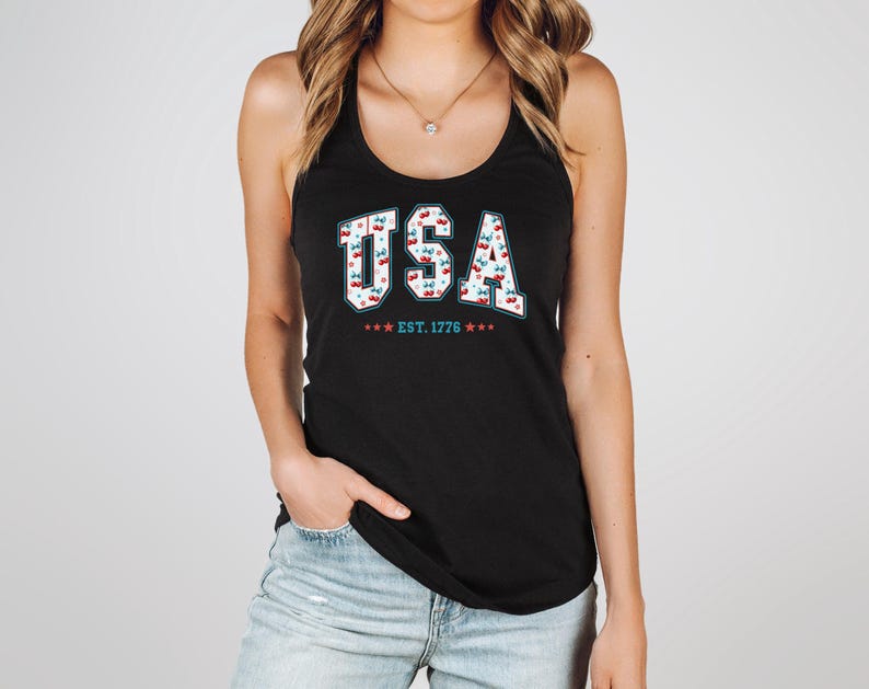 May include: Black tank top with the word "USA" in white, outlined in red, and filled with a cherry pattern. Below the word "USA" is the text "EST. 1776" with red and blue stars. The tank top is worn by a person.