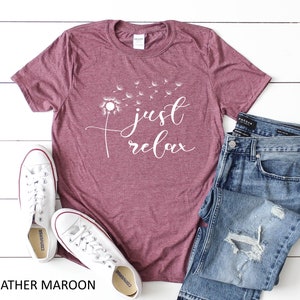 May include: Heather maroon t-shirt with a white dandelion graphic and the text "just relax". The shirt is laid flat on a white surface with a pair of white sneakers and blue jeans.