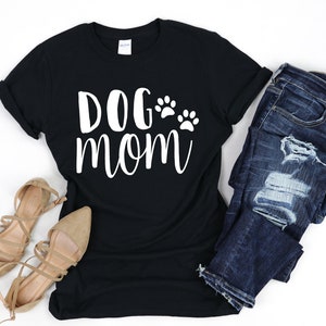 May include: Black t-shirt with the text "DOG mom" in white script, accompanied by paw print graphics. The shirt is paired with blue jeans and tan flats, creating a casual outfit.