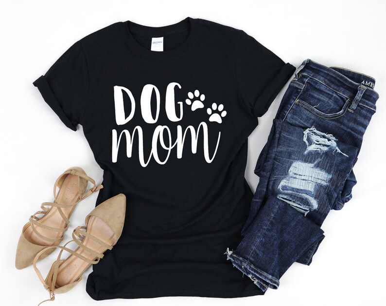 May include: Black t-shirt with the text "DOG mom" in white script, accompanied by paw print graphics. The shirt is paired with blue jeans and tan flats, creating a casual outfit.
