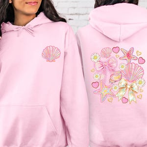 May include: Pale pink hoodie featuring a seashell motif on the front and a larger design on the back. The design includes seashells, bows, hearts, and stars in pastel shades.