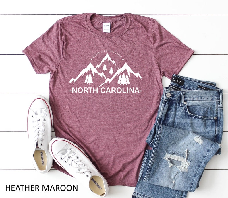 May include: Heather maroon t-shirt with a white graphic of mountains and trees with the text "Never Stop Exploring - North Carolina -" printed on the front. The t-shirt is laid flat on a white surface with a pair of white sneakers and blue jeans.