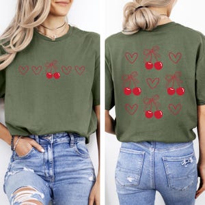 May include: A green t-shirt with a cherry and heart design on the front and back. The front design has a row of cherries with bows and hearts. The back design has multiple cherries with bows and hearts.