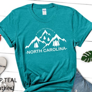 May include: Teal heather t-shirt with a white graphic of mountains and trees. The text "Never Stop Exploring - North Carolina" is printed on the shirt.