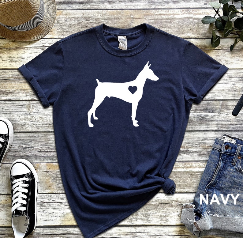 May include: A navy blue t-shirt with a white silhouette of a Doberman Pinscher with a heart in its chest. The shirt is made of a soft, comfortable material and is perfect for everyday wear.