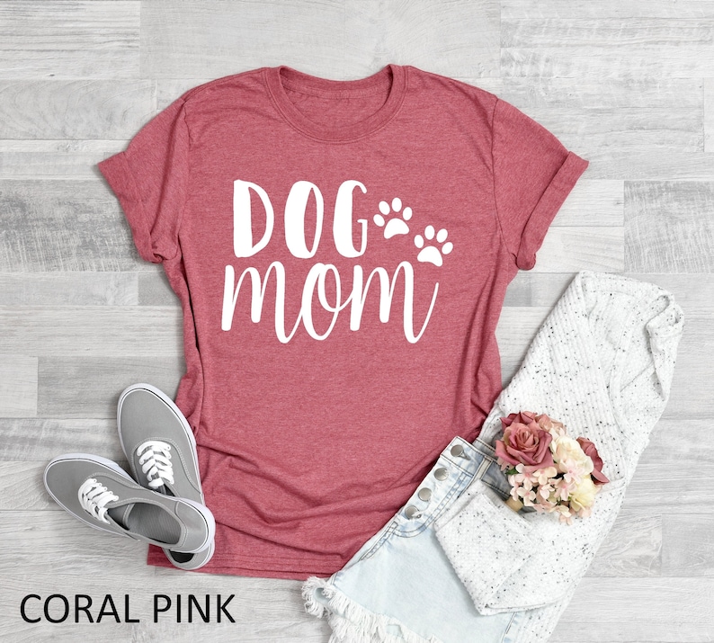May include: Coral pink t-shirt with the words "DOG MOM" in white script and two paw prints. The shirt is paired with gray sneakers, denim shorts, and a white sweater. A bouquet of pink flowers rests nearby.