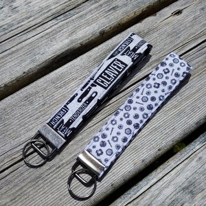 May include: Two keychains with silver key rings. One keychain has a white and black fabric strap with the words "Meat Cleaver" and "Tenderizer" printed on it. The other keychain has a white and black fabric strap with a pattern of screws and nuts.