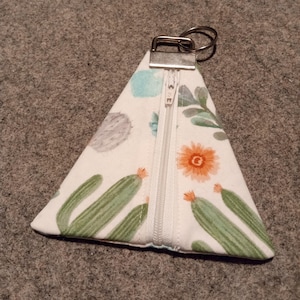 May include: A white triangular zippered pouch with a cactus print. The pouch has a silver zipper and a silver ring for attaching keys.