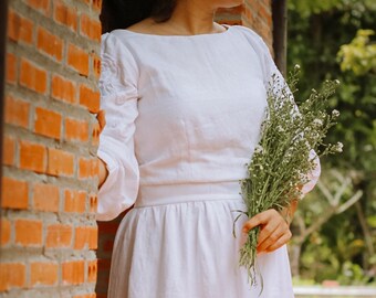 May include: A woman in a white linen dress holds a bouquet of white flowers. The dress has a simple, elegant design with a boat neckline and long sleeves. The woman is standing in front of a brick wall.