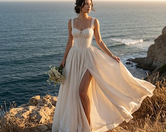 Puede incluir: Un vestido de novia color crema con una abertura alta y un corpiño ajustado. El vestido tiene un escote corazón y tirantes finos. La modelo sostiene un ramo de flores blancas y está de pie en un acantilado con vistas al océano.