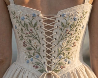 May include: A beige corset-style bodice featuring intricate floral embroidery with white, blue, and pink flowers and green leaves. The back includes a central lace-up closure with matching beige cord threaded through silver-toned metal eyelets.