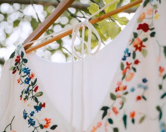 May include: A white dress with colorful floral embroidery hangs on a wooden hanger. The embroidery features red, blue, orange, and green flowers and leaves. The dress has a V-neck and tie detail at the back.