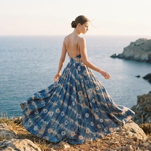 May include: A flowing, floor-length dress with a blue and white tie-dye pattern. The dress has thin straps and a low back. The image is taken outdoors, with a rocky cliff and ocean in the background. The dress is in motion.