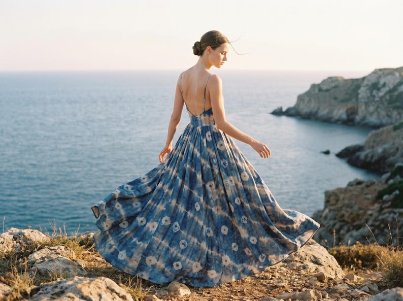 May include: A flowing, floor-length dress with a blue and white tie-dye pattern. The dress has thin straps and a low back. The image is taken outdoors, with a rocky cliff and ocean in the background. The dress is in motion.