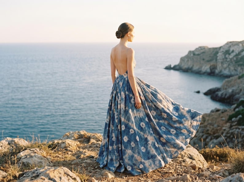 May include: A flowing, backless blue and white patterned dress. The dress is worn by a person standing on a rocky cliff overlooking the ocean. The dress's fabric billows in the wind, creating a dramatic effect. The scene is bathed in soft sunlight.