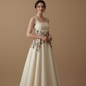 May include: An ivory-colored sleeveless gown with a square neckline and a full skirt. The dress features floral embroidery in shades of purple, yellow, and green around the waist and cascading down the skirt. The dress is made of a textured fabric.