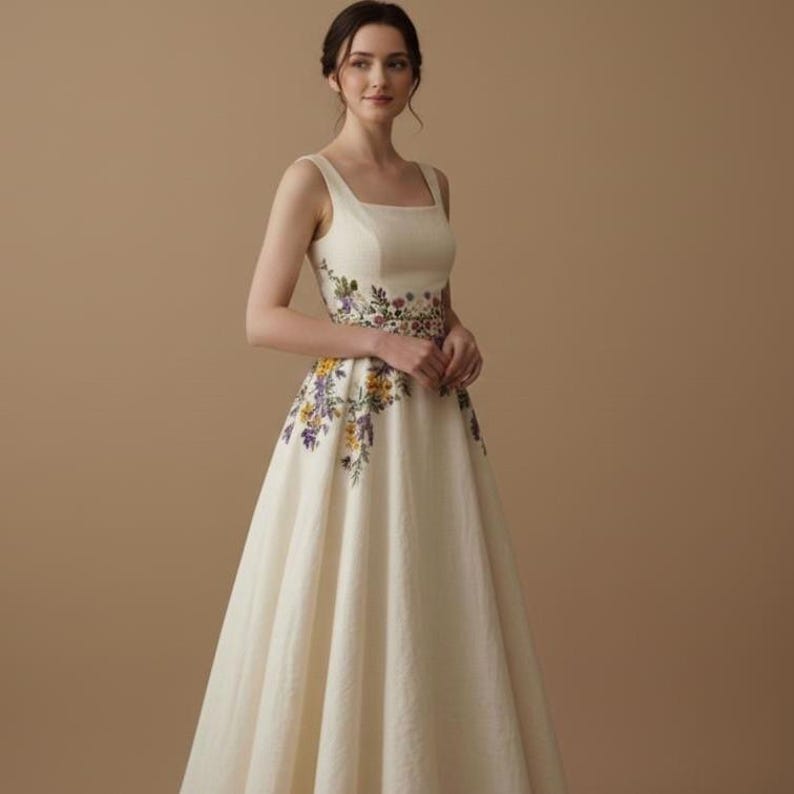 May include: An ivory-colored sleeveless gown with a square neckline and a full skirt. The dress features floral embroidery in shades of purple, yellow, and green around the waist and cascading down the skirt. The dress is made of a textured fabric.