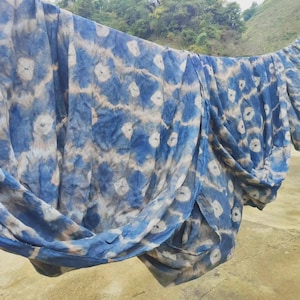 May include: Blue and white tie-dye fabric hanging outdoors. The fabric has a repeating pattern of white circles against a blue background, with areas of tan and blue. The fabric is draped over a line, with a natural landscape in the background.