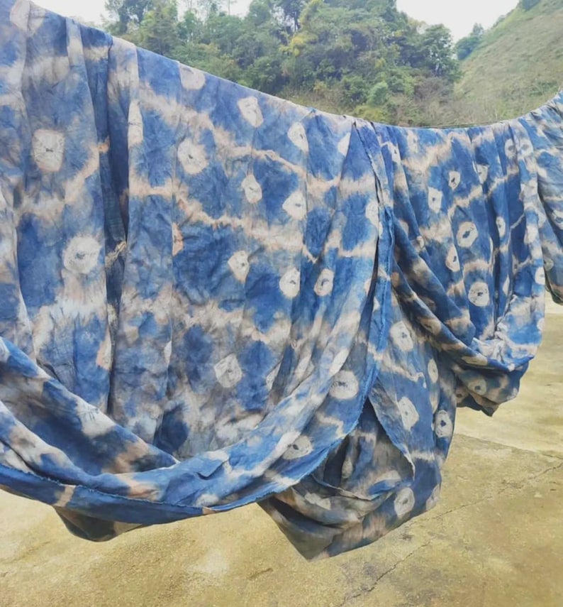 May include: Blue and white tie-dye fabric hanging outdoors. The fabric has a repeating pattern of white circles against a blue background, with areas of tan and blue. The fabric is draped over a line, with a natural landscape in the background.
