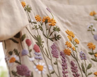 May include: A beige linen dress with embroidered wildflowers in shades of yellow, purple, pink, and white. The floral embroidery is detailed and covers the bodice of the dress, creating a delicate, handcrafted look.