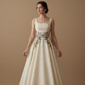 May include: A cream-colored sleeveless dress with a square neckline and a full skirt. The waistline is adorned with a colorful floral embroidery featuring purple, yellow, and green flowers. The dress is made of a smooth, flowing fabric.