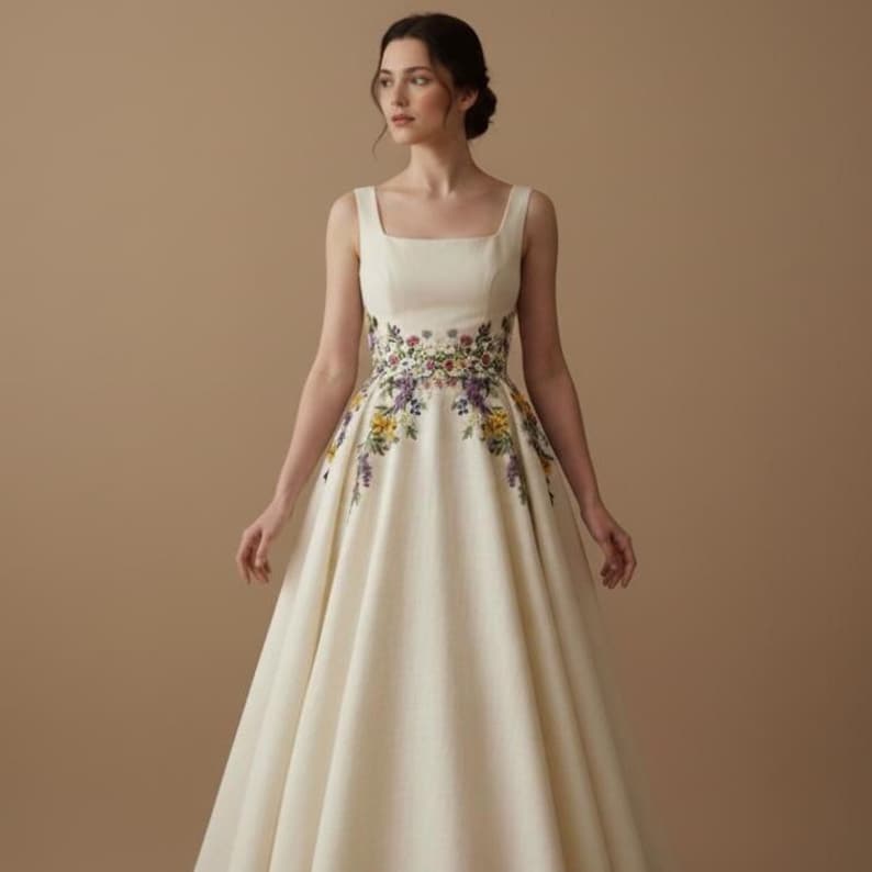 May include: A cream-colored sleeveless dress with a square neckline and a full skirt. The waistline is adorned with a colorful floral embroidery featuring purple, yellow, and green flowers. The dress is made of a smooth, flowing fabric.