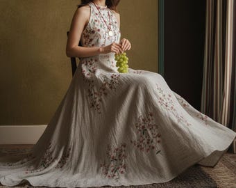 May include: A light-colored, floor-length dress with floral embroidery. The dress features a halter neckline and a flowing skirt. The person is holding a bunch of green grapes. The dress is worn while seated on a wooden chair.