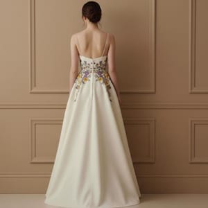 May include: Ivory-colored formal gown with floral embroidery. The dress features a fitted bodice with thin straps and a full, flowing skirt. The back of the dress is adorned with colorful floral embroidery in yellow, purple, and green.