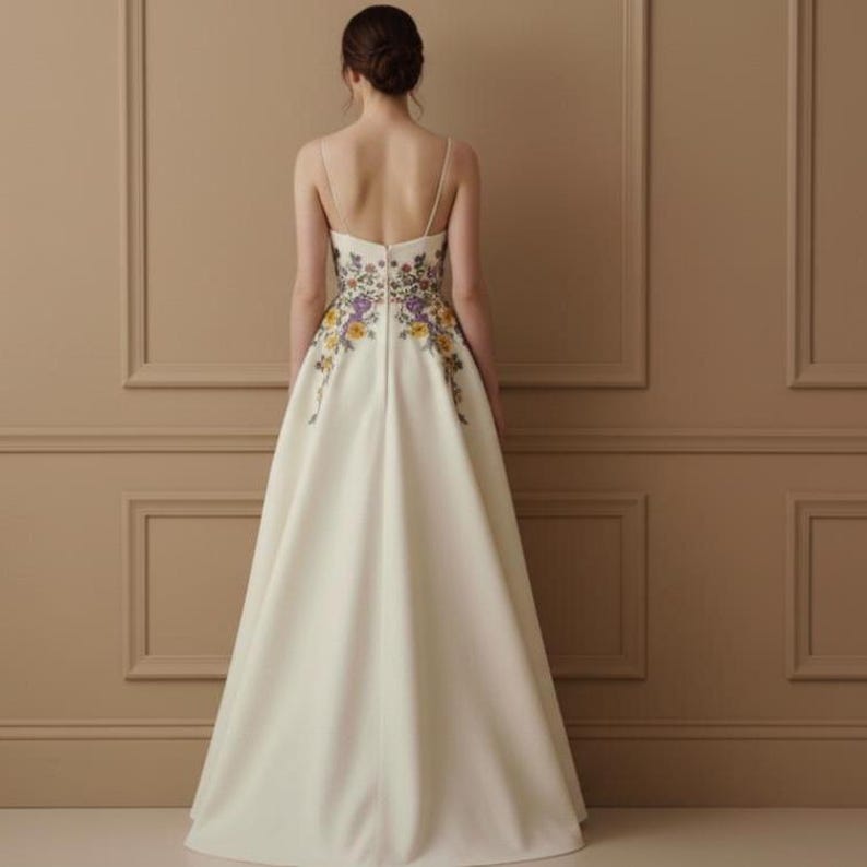 May include: Ivory-colored formal gown with floral embroidery. The dress features a fitted bodice with thin straps and a full, flowing skirt. The back of the dress is adorned with colorful floral embroidery in yellow, purple, and green.