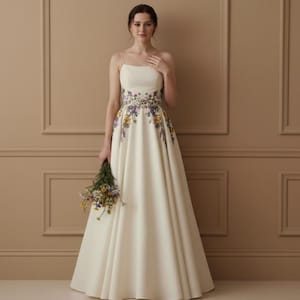May include: An ivory-colored wedding dress with a floral embroidered waistline and spaghetti straps. The dress has a flowing A-line skirt and is paired with a bouquet of wildflowers. The dress is a classic and elegant choice for a wedding.