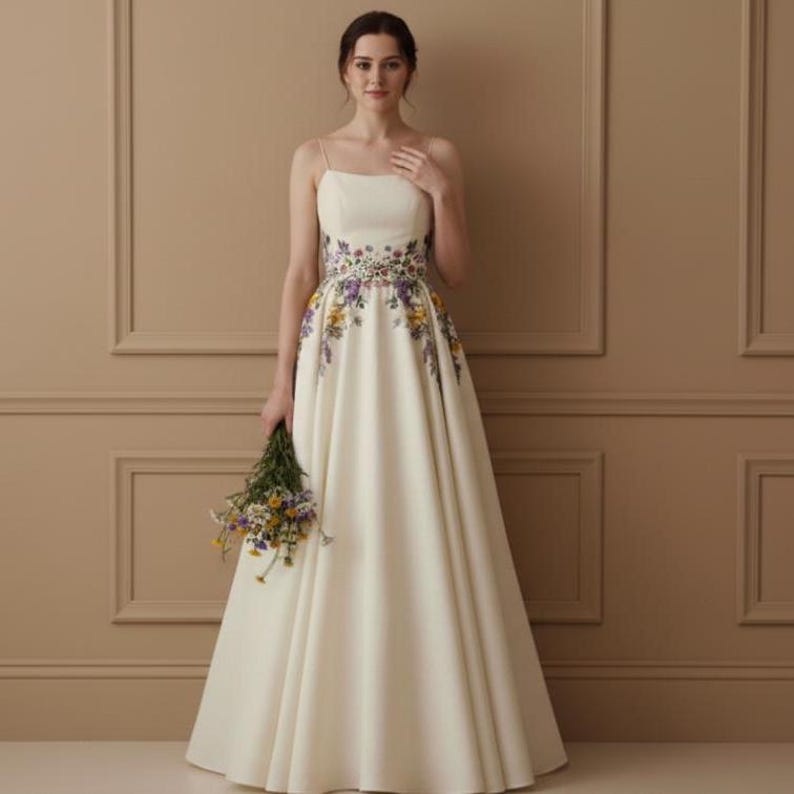 May include: An ivory-colored wedding dress with a floral embroidered waistline and spaghetti straps. The dress has a flowing A-line skirt and is paired with a bouquet of wildflowers. The dress is a classic and elegant choice for a wedding.