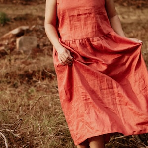 Könnte beinhalten: Eine Frau trägt ein langes, fließendes Leinenkleid in Korallenfarbe. Das Kleid hat einen gerafften Rock und ein schlichtes, ärmelloses Oberteil. Die Frau steht auf einem Feld mit trockenem Gras und Ästen.