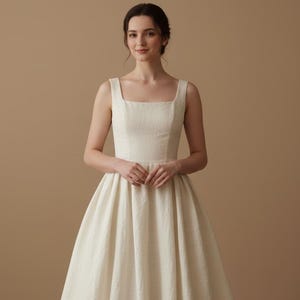May include: A cream-colored sleeveless dress with a square neckline and a full skirt. The dress is made of a textured fabric and is worn by a person. The background is a solid tan color.
