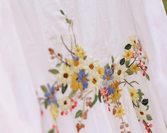May include: White fabric with embroidered floral design. The embroidery features yellow, blue, and white flowers with green leaves and red berries. The fabric appears to be a light, flowing material.