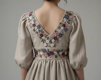May include: A beige dress with a V-neckline and elbow-length sleeves. The dress features floral embroidery in shades of red, purple, blue, and white along the neckline and waist. The dress has a gathered skirt.