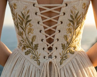 Puede incluir: Vestido color crema con corpiño estilo corsé y espalda con cordones. El corpiño presenta intrincados bordados florales en tonos dorados y verdes. El vestido tiene un escote cuadrado y tirantes anchos.