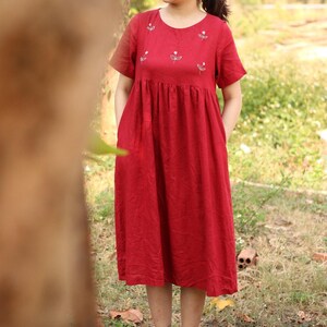 May include: A red linen dress with white embroidered flowers. The dress has a round neckline and short sleeves. It is a midi length dress with a gathered waist.