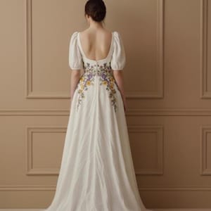 May include: A white wedding dress with a floral embroidered design in yellow and purple. The dress features short, puffy sleeves and a low back. The dress is floor-length and has a flowing skirt.