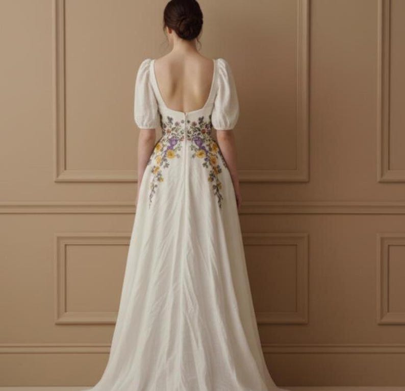May include: A white wedding dress with a floral embroidered design in yellow and purple. The dress features short, puffy sleeves and a low back. The dress is floor-length and has a flowing skirt.