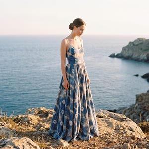 May include: A long, flowing blue dress with a white pattern. The dress has thin straps and a full skirt. The model is standing on a rocky cliff overlooking the ocean. The dress is made of a lightweight fabric, perfect for warm weather.