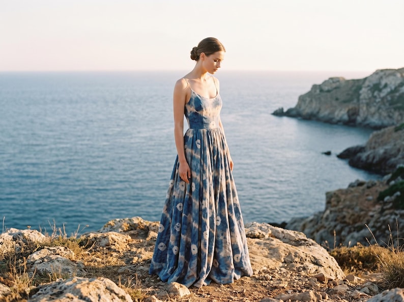 May include: A long, flowing blue dress with a white pattern. The dress has thin straps and a full skirt. The model is standing on a rocky cliff overlooking the ocean. The dress is made of a lightweight fabric, perfect for warm weather.