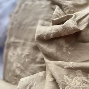 Pode incluir: Close-up de tecido bege com bordado floral branco. O tecido é drapeado, mostrando dobras e texturas. O bordado apresenta padrões florais e de folhas detalhados, criando um design delicado e elegante. A estética geral é suave e natural.