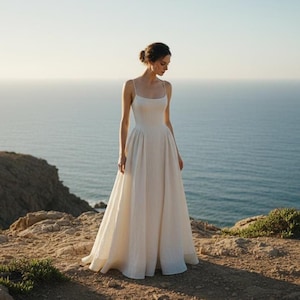 Könnte beinhalten: Ein weißes Hochzeitskleid mit einem figurbetonten Oberteil und einem ausgestellten A-Linien-Rock. Das Kleid hat dünne Spaghettiträger und ein schlichtes, elegantes Design. Die Frau steht auf einer felsigen Klippe mit Blick auf das Meer.