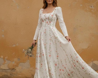May include: A white, long-sleeved wedding dress with a square neckline and floral embroidery. The dress features a flowing skirt and is adorned with delicate pink flowers and green stems. The woman is holding a small bouquet of flowers.