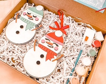 Pode incluir: Um kit de artesanato com dois enfeites de boneco de neve de madeira, tinta, pincéis e cola. O kit inclui instruções que dizem "Você quer ser um boneco de neve?" e "Feliz Natal!"
