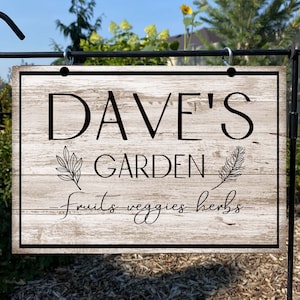 Puede incluir: Un letrero de madera rústico con el texto "Dave's Garden - fruits - veggies - herbs" en letras negras. El letrero está colgado de un gancho de metal negro.