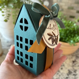 Könnte beinhalten: Eine teal-farbene, hausförmige Geschenkbox mit ausgeschnittenen Fenstern und einem runden Fenster. Sie ist mit einem grünen Samtband und einem Holzanhänger verziert. Der Anhänger trägt den Namen "Michelle" und ein Tannenzapfen-Design.